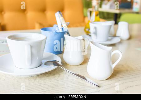 Service de petit déjeuner. Café, lait et sucre dans un ustensile blanc. Tasse à café et pichet à lait sur la table dans le café. Intérieur du café. Vaisselle blanche. Banque D'Images