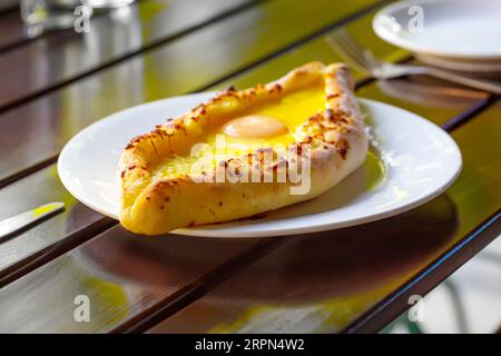 Le khachapuri de style Adjara, pâte cuite au four avec œuf, fromage et deux morceaux de beurre sur une assiette blanche dans un café. Banque D'Images