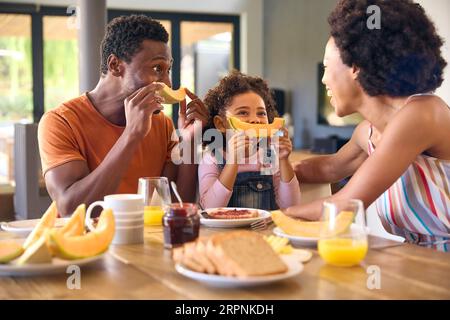 Photo en famille avec parents et fille à la maison faire des visages drôles avec Melon avant de manger petit déjeuner Banque D'Images