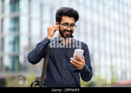 Souriant jeune homme indien portant des écouteurs tenant le téléphone tout en marchant Banque D'Images