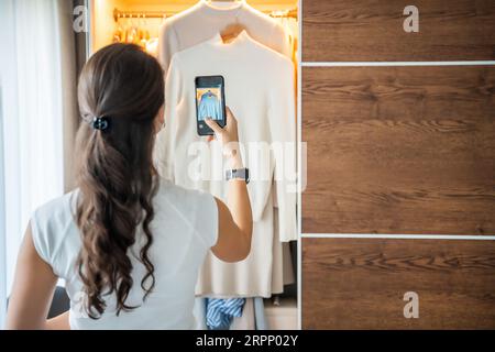 Jeune femme prenant des photos de vieux vêtements indésirables à vendre dans l'application smartphone Banque D'Images