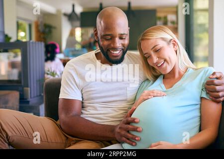 Aimer couple multi-racial avec femme enceinte sur canapé à la maison avec famille multi-génération derrière Banque D'Images