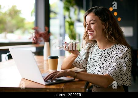 Femme d'affaires avec café travaillant sur ordinateur portable dans café parler dans le microphone du téléphone portable Banque D'Images