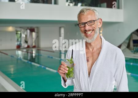 concept de retraite de bien-être, homme mûr joyeux en robe blanche tenant un cocktail près de la piscine dans le centre thermal Banque D'Images