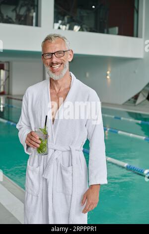 concept de retraite de bien-être, homme mûr heureux en robe blanche tenant un cocktail près de la piscine dans le centre de spa Banque D'Images