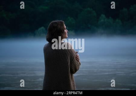 Une femme en chandail sur la rive d'une rivière brumeuse dans la soirée. Banque D'Images