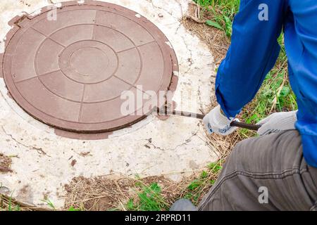 Un plombier ouvre un couvercle de trou d'homme sur un puits de béton avec un pied de biche. Inspection et entretien des puits d'eau et d'égout. Banque D'Images