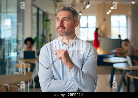 Portrait de souriant homme d'affaires mature tenant des lunettes debout dans le bureau occupé Banque D'Images