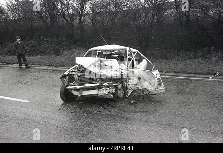 1983, historique, un homme debout sur le bord d'une route principale regardant sa voiture gravement écrasée, ayant été impliqué dans une collision frontale sur une route humide et glissante de landes, Angleterre, Royaume-Uni. Banque D'Images