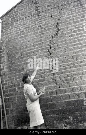 1983, historique, chalet de carrière, une dame devant sa maison tenant un morceau de rocher et pointant vers une grande fissure verticale dans le mur latéral, due à l'affaissement se produisant lorsque le sol glisse, car il a été construit près d'une carrière, Yorkshire, Angleterre, Royaume-Uni. Banque D'Images