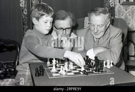 1980, assis avec deux hommes plus âgés, dont l'un est son grand-père, un jeune garçon jouant une partie d'échecs avec eux, Angleterre, Royaume-Uni, le garçon frappe sur la tour de l'homme plus âgé. Les échecs sont un jeu de société populaire d'habileté stratégique impliquant deux joueurs, dans le but de rendre impossible l'évasion du roi de l'adversaire. Banque D'Images