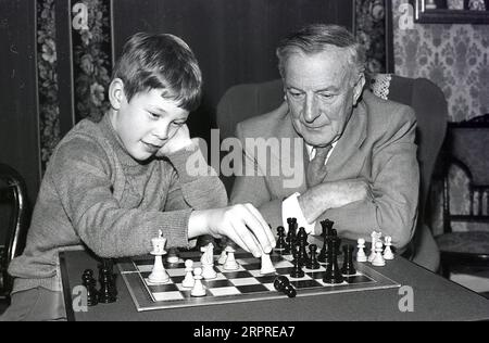 1980, assis avec son grand-père à l'intérieur d'un front rom un jeune garçon jouant une partie d'échecs avec lui, Angleterre, Royaume-Uni. Les échecs sont un jeu de société populaire d'habileté stratégique impliquant deux joueurs, dans le but de rendre impossible l'évasion du roi de l'adversaire. Banque D'Images