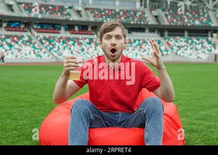 Fan de football choqué en tshirt rouge avec verre de bière regardant le match de football dans la zone des fans du stade. Banque D'Images
