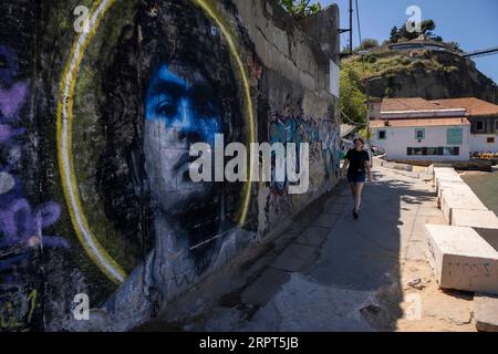 Une femme vue marchant près d'un mur avec l'image du joueur de football Diego Maradona, dans la ville d'Almada. Situé sur la rive sud du fleuve Tejo, Almada est le meilleur point de vue sur la ville de Lisbonne, avec le château, l'ascenseur panoramique de Boca do Vento et la statue du Christ Roi construite en 1959, ils sont utilisés comme points d'observation. Banque D'Images