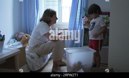 Scène franche de grand-mère interagissant avec petit-fils enfant dans la chambre à coucher regardant des jouets et des objets et conversant Banque D'Images