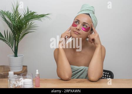 Jeune femme réfléchie avec des patchs sous les yeux réutilisables assis à la procédure de soin de la dame de table Banque D'Images