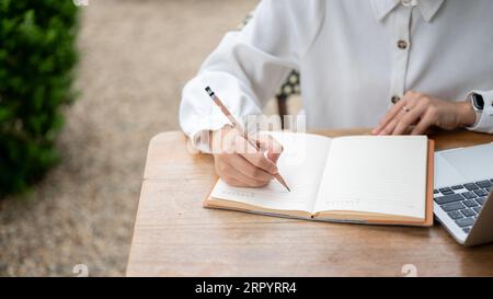 Gros plan image de dessus d'une femme écrivant quelque chose dans son carnet, tenant un journal ou faisant ses devoirs tout en se relaxant à une table à l'extérieur. Banque D'Images