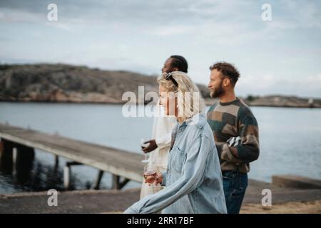 Vue latérale de la jeune femme appréciant des boissons tout en marchant avec des amis près du lac Banque D'Images