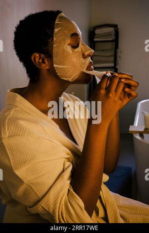 Jeune femme enlevant le masque facial sur le visage tout en étant assise dans la salle de bain Banque D'Images