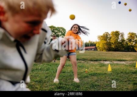 Fille souriante lançant la balle sur un ami tout en jouant dans le terrain de jeu au camp d'été Banque D'Images