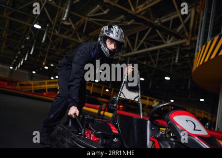 motorsport et speed drive, pilote de kart concentré en casque et sportswear poussant le kart sur circuit Banque D'Images