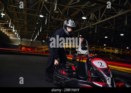 motorsport et speed drive, pilote de kart concentré en sportswear et casque poussant le kart sur circuit Banque D'Images