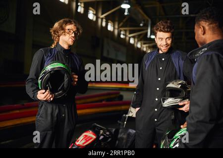 hommes interraciaux positifs discutant à l'intérieur du circuit de kart, trois coureurs de kart tenant des casques Banque D'Images