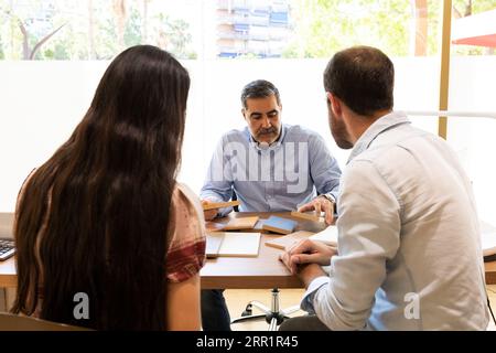Vue arrière couple anonyme regardant vers le bas tout en étant assis à la table du magasin de meubles avec le mâle et vérifiant des morceaux d'échantillon en bois à la lumière du jour Banque D'Images