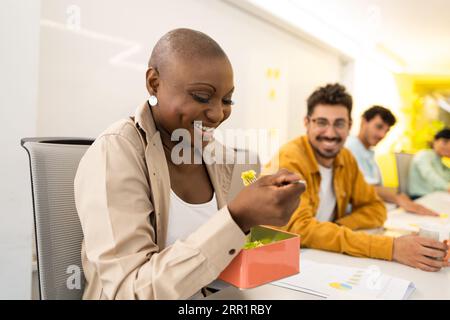 Du dessous de la femme noire heureuse regardant vers le bas avec le plat dans la cuillère de la boîte tout en étant assis avec un mâle souriant flou regardant la caméra dans les lumières de bureau Banque D'Images