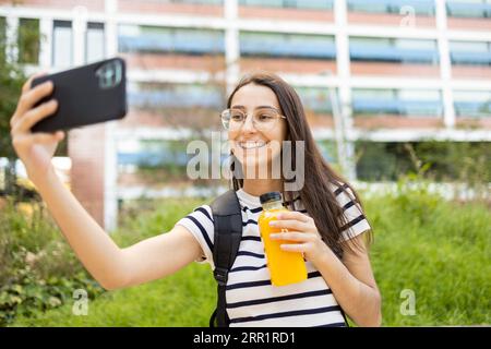 Heureuse jeune femme dans des lunettes avec sac à dos et bouteille de jus frais prenant selfie sur la rue de la ville tout en souriant joyeusement Banque D'Images