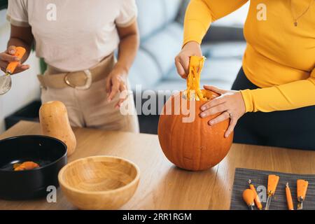 Amis féminins anonymes debout à table avec des outils d'Halloween et tirant la pulpe du dessus de la citrouille orange à la lumière du jour Banque D'Images