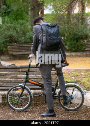 Vue arrière d'un homme anonyme en costume formel avec sac à dos regardant vers le bas tout en se tenant dans le parc et avec chaussure jambe parking vélo sur stand en plein jour Banque D'Images