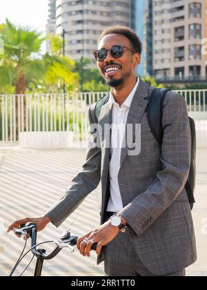 Mec afro-américain souriant en tenue formelle et lunettes de soleil debout avec vélo contre le bâtiment moderne et regardant loin Banque D'Images