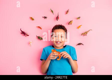 Vue d'angle de dessus photo de joli petit garçon mignon porter un t-shirt bleu jouant des jouets dino préférés fond isolé de couleur rose Banque D'Images