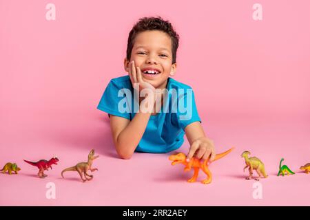 Photo de mignon petit garçon rêveur porter un t-shirt bleu jouant des jouets dinosaures fond isolé de couleur rose Banque D'Images