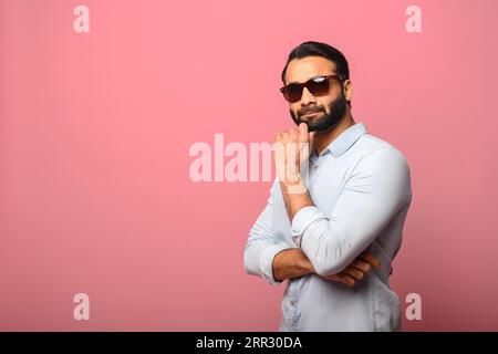 Bel homme barbu indien d'âge moyen portant des lunettes de soleil reposant le menton sur la main regardant la caméra avec réflexion, pensant aux vacances debout sur fond rose Banque D'Images