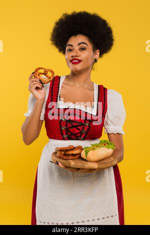 joyeuse serveuse afro-américaine de l'oktoberfest tenant des bretzels et hot-dog sur un plateau en bois sur jaune Banque D'Images