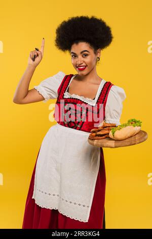 joyeuse serveuse afro-américaine oktoberfest avec des collations sur un plateau en bois montrant le signe d'idée sur jaune Banque D'Images