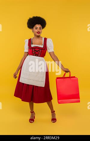 heureuse serveuse bavaroise afro-américaine dans la tenue de l'oktoberfest tenant des sacs à provisions sur jaune Banque D'Images