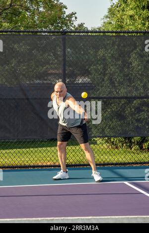 Un joueur de pickleball masculin retourne un service d'une balle jaune vif avec son revers sur un terrain dédié dans un parc public. Banque D'Images