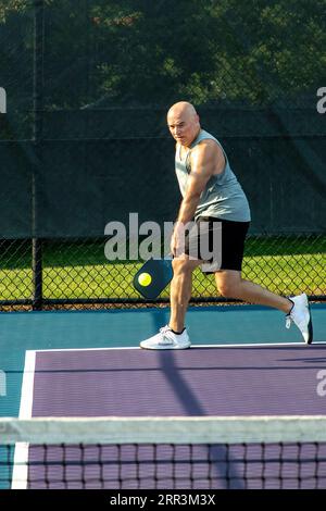 Un joueur de pickleball masculin retourne un service d'une balle jaune vif avec son revers sur un terrain dédié dans un parc public. Banque D'Images