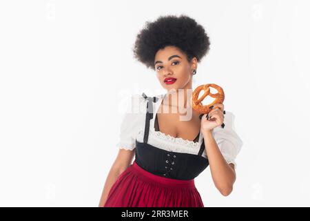 jolie serveuse bavaroise afro-américaine en tenue oktoberfest tenant délicieux bretzel sur blanc Banque D'Images