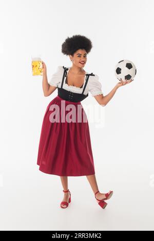 serveuse afro-américaine oktoberfest avec tasse de bière mousseuse jouant au football sur blanc, pleine longueur Banque D'Images