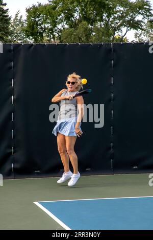 Une joueuse de pickleball retourne un service d'une balle jaune vif sur un terrain dédié dans un parc public. Banque D'Images