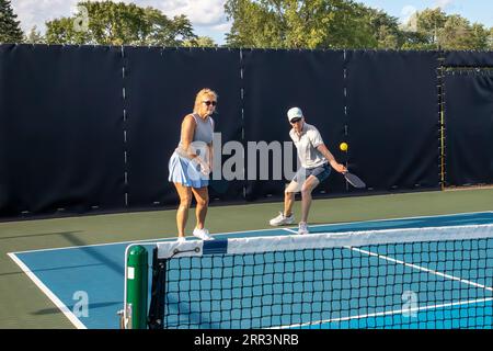 Un joueur de pickleball masculin renvoie une balle alors que sa partenaire féminine se tient près de la ligne de base. Banque D'Images