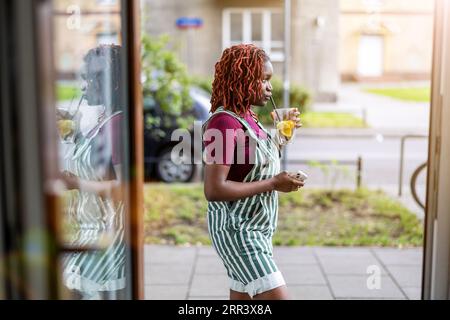 Jeune personne non binaire buvant du jus debout dans l'embrasure d'un café Banque D'Images