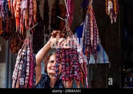 Une vendeuse coupe des brins de churchkhela (géorgien ჩურჩხელა) à son étal au Dezerter Bazaar (Marché des déserteurs) à Tbilissi, en Géorgie. Les bras levés, elle tient un paquet et une paire de ciseaux et regarde les brins avec un sourire. Les brins en rouge, violet, orange et blanc pendent en grappes denses sur des cordes. La churchkhela est une confiserie géorgienne. Les noix sont enrobées de pelamushi, un sirop de jus de raisin réduit avec de l'amidon et sans sucre. Pour la préparer, des noix de walnoute ou des noisettes sont enfilées sur une corde, trempées plusieurs fois dans du pelamushi puis séchées. La churchkhela est une provision classique pour Banque D'Images