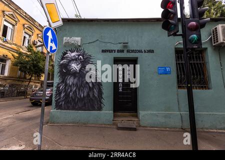 Raven's Bar (géorgien რეივენს ბარი), un bar à cocktails dans le quartier de Chugureti à Tbilissi, en Géorgie, possède une fresque murale noir et blanc d’un corbeau ébouriffé sur sa façade enduite de vert turquoise. Le nom apparaît en anglais et en géorgien au-dessus de la porte noire et verticalement sur la porte elle-même. Un panneau bleu indiquant le numéro de la maison montre le numéro 11. Un feu de circulation et un panneau de signalisation se trouvent devant. Un bâtiment résidentiel enduit de jaune se dresse à gauche. Banque D'Images