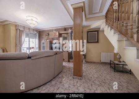 Un salon avec des meubles en bois, des murs de couleur crème, des lambris en bois, des escaliers avec une balustrade, des sols en grès et un pilier au milieu Banque D'Images