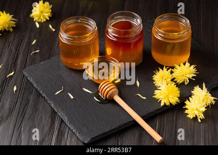 Une variété de miel dans des pots, une pelle et des fleurs sur une plaque de pierre noire sur un fond en bois. Le concept de produits apicoles biologiques. Banque D'Images